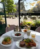 A high-quality, well-composed photograph showcasing a breakfast spread at The Ivy. The image features a table filled with various breakfast items, including pastries, smoked salmon, and drinks, set against a scenic backdrop of the Exeter Cathedral visible through the window. The natural lighting and professional styling effectively convey an upscale, inviting dining experience.