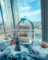 A high-angle shot of an afternoon tea service featuring a tiered stand, cocktails, and dry ice, set against a panoramic London skyline view.