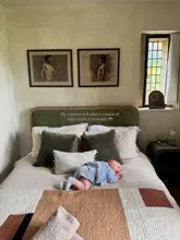 A lifestyle-focused, candid shot taken from a 'camera roll' perspective, featuring a baby lying on a bed in a Cotswolds accommodation. The aesthetic is 'cottage core' with neutral tones, soft natural lighting, and a relaxed, homely atmosphere.