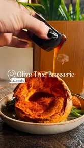 A short, polished promotional video for Olive Tree Brasserie in Leeds, focusing on their Sunday roast sharing board. The video features close-up shots of food preparation, specifically gravy being poured over a Yorkshire pudding, followed by shots of the sharing platter and cocktails in a nicely lit restaurant setting.