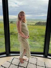 A portrait of a pregnant woman standing in front of a large, floor-to-ceiling window at a glamping venue, showcasing the scenic countryside view.