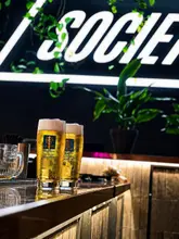 A high-quality, atmospheric shot of two glasses of Augustiner lager served at a bar, highlighting the venue's focus on premium beer and a lively, sports-friendly environment.