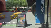 A static shot of the exterior of the OXO Gallery, featuring an A-board sign advertising a live sand mandala creation event. The image captures the entrance area of the venue on a sunny day.