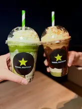 A high-quality, studio-style shot featuring two hands holding up plastic cups of matcha-based drinks against a dark, minimalist background, effectively highlighting the product branding.