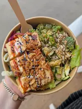 A vibrant, top-down shot of a healthy salad bowl featuring satay tofu, kimchi, and crispy onions, held in a hand against a casual market background.