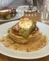 A close-up, eye-level shot of a stack of buttermilk pancakes topped with stewed rhubarb, a dollop of mascarpone, and almond crumb. The image is styled for a brunch review, featuring text overlay that highlights the dish.