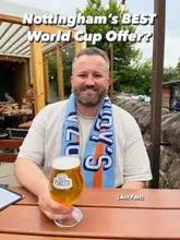 A promotional image featuring a man holding a pint of beer at an outdoor venue, highlighting a World Cup-themed promotional offer involving a branded scarf. The image is well-composed, using natural lighting and a shallow depth of field to keep the focus on the subject and the product.