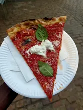 A high-quality, top-down shot of a single slice of pizza topped with fresh basil and a dollop of cheese, held on a paper plate against a blurred outdoor background.