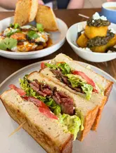 A bright, natural-light food photograph capturing a cafe-style brunch spread. The focus is on a sandwich in the foreground, with other dishes like eggs and potatoes visible in the background, creating a warm and inviting atmosphere.