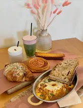 A high-quality, well-lit flat lay photograph showcasing a variety of brunch items from a bakery, including a tomato danish, a turkey croissant, baked eggs, and specialty drinks.
