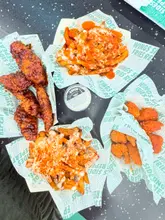 A top-down, flat-lay photograph of a fast-food meal from Wingstop, featuring baskets of chicken tenders, boneless bites, and loaded fries, arranged around a central pot of ranch dip on a dark table.