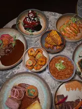 A top-down shot of a table spread featuring various Afro-Caribbean dishes, including wings, grilled corn, shrimp, rice, and steak, served in a dimly lit restaurant setting.