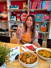 A photo of a woman holding a glass of wine at a table with two Asian dishes, set against a backdrop of a retail shelf filled with cookbooks and kitchenware at a cooking school.