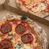 A close-up, high-angle shot of two pizzas in cardboard takeaway boxes, featuring a chorizo and rosemary pizza in the foreground and a margherita in the background.