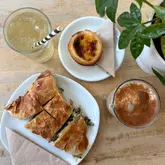A bright, overhead flat-lay shot of a cafe lunch spread, featuring a spinach and feta pie, a pastel de nata, and two iced drinks. The natural lighting creates an inviting, fresh atmosphere suitable for a casual cafe setting.