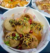 A close-up, top-down shot of a bowl of Indian street food, specifically gol gappe, served in a branded paper-lined bowl. The image focuses on the textures and toppings of the dish.