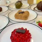 A high-quality, close-up shot of a red, glazed dessert from Haute Dolci, with other patisserie items blurred in the background, showcasing the brand's new range.