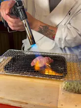 A high-quality, dramatic close-up shot capturing a chef blow-torching a piece of sushi on a grill, highlighting the premium nature of the omakase experience.