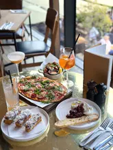 A high-angle, vibrant shot of a table spread at The Lighterman, featuring a variety of dishes including pizza, burrata, bruschetta, and chicken wings, alongside cocktails. The setting is bright and airy, capturing a summer dining atmosphere.