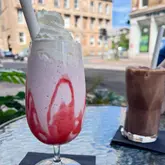 A casual, eye-level shot of a strawberry milkshake served in a tall glass with whipped cream, positioned on an outdoor table at a cafe. A chocolate drink is visible in the background, with a street scene behind the table.