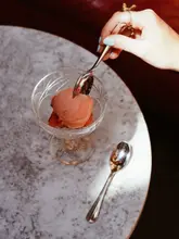 A close-up, top-down shot of a single scoop of pink sorbet served in a crystal coupe glass on a marble table, with a silver spoon resting beside it. The lighting is soft and warm, highlighting the texture of the sorbet and the elegant glassware, creating an inviting, summery atmosphere.