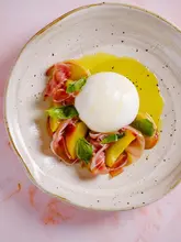 A high-quality, professional overhead food photograph featuring a burrata salad with roasted peaches, prosciutto di parma, and fresh basil, drizzled with olive oil on a textured ceramic plate.