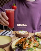 A vibrant, well-composed shot of a Greek dining spread featuring a strawberry margarita, a feta-topped dip, and grilled shrimp, with a person in a purple shirt softly blurred in the background.