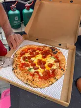 A high-angle shot of a freshly baked Neapolitan-style pepperoni pizza in a cardboard box, being sliced by a pizza cutter. The setting appears to be a casual outdoor pop-up environment, evidenced by the gas canisters in the background.