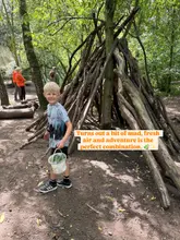 A bright, natural photograph of a young boy holding a bucket in a woodland setting, standing next to a stick-built den. The image promotes an outdoor holiday club for children, capturing a moment of play and exploration in nature.
