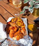 A top-down, close-up shot of a panko chicken spice bag served in foil, accompanied by a pot of curry sauce and a glass of white wine, set on a wooden outdoor table.
