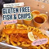 A bright, well-lit, top-down shot of a takeaway box containing gluten-free fish and chips, accompanied by pots of curry sauce and mushy peas, set against a beach backdrop.