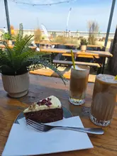 A casual, sunlit shot of a slice of carrot cake and two iced coffees served on a wooden table at an outdoor venue. The image captures a relaxed, coastal cafe atmosphere, with the food and drinks positioned in the foreground against a bright, open-air background.