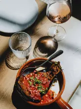 A high-quality, top-down shot of a Lebanese fattet aubergine dish served in a rustic ceramic bowl, accompanied by a glass of rosé wine on a wooden table. The lighting is soft and natural, highlighting the textures of the tomato sauce, pomegranate seeds, and fresh herbs.