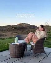 A woman sits in a wicker chair on a patio overlooking a scenic Scottish landscape during golden hour, holding a mug. The image conveys a sense of relaxation and escape, promoting a luxury stay on the Isle of Luing.