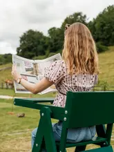 A lifestyle photograph featuring a person sitting on a green wooden chair in a countryside setting, reading a newspaper that mentions the venue. The image conveys a relaxed, peaceful atmosphere suitable for a hotel or estate promotion.