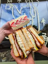 A high-quality, first-person perspective shot showcasing three Japanese-style sandwiches held up against a cafe window. The image highlights the cross-sections of a fruit sando and two katsu sandos, emphasizing the fresh ingredients and textures.