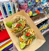 A close-up, eye-level shot of two tacos served in a cardboard tray, held by a person. The tacos are topped with fresh slaw, herbs, and chili slices. The background is a vibrant but highly distracting wall of fabric rolls in a market setting.