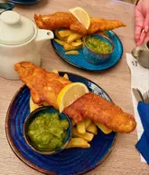 A casual, overhead-style shot of two plates of fish and chips served in a garden center cafe. The food is presented on blue ceramic plates with a side of mushy peas and a lemon wedge. A teapot and a receipt are also visible on the table.