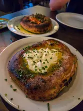 A close-up, eye-level shot of two rustic flatbreads topped with melted cheese, herbs, and oil, served on a dark wooden table in a restaurant setting.