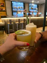 A first-person perspective shot of two people toasting with cocktails in a bar setting. The foreground drink is a yellow cocktail garnished with a kiwi slice, while the background drink is a creamy, tropical-style cocktail with a pineapple leaf. The background shows a bar counter with stools and ambient lighting.