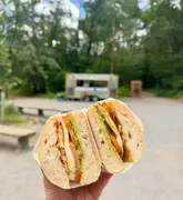 A close-up, eye-level shot of a freshly prepared caprese sandwich held in front of a food trailer located in a park setting. The image effectively highlights the product while using a shallow depth of field to provide context about the venue's outdoor location.
