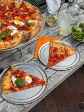A high-quality, well-composed shot of two slices of pizza served on metal trays on a rustic wooden table, accompanied by drinks and a menu, effectively showcasing the venue's casual dining atmosphere.