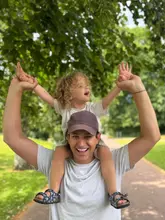 A high-quality, candid-style portrait of a father carrying his child on his shoulders in an outdoor setting, likely a park or attraction. The image captures a joyful family moment with soft, natural lighting and a pleasant background blur, effectively conveying a positive, family-friendly atmosphere.