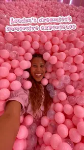 A bright, selfie-style shot of a creator immersed in a pink ball pit at the House of Dreamers exhibition in London. The image effectively communicates the fun, interactive nature of the venue.
