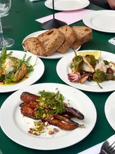 A flat-lay photograph of a shared meal on a green table, featuring sourdough bread, a salad with figs and cheese, grilled squid with peppers, and roasted carrots with pistachios.