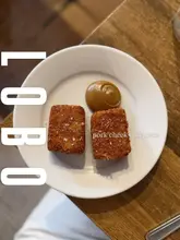 An overhead, top-down shot of two pork cheek croquettes served on a white plate with a dollop of sauce, set against a wooden table. The image features prominent, large white text overlays identifying the venue and the dish.