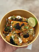 A high-quality, close-up overhead shot of hash browns topped with caviar, chives, and chili flakes, served in a disposable paper bowl. The lighting is soft and natural, highlighting the textures of the food, and the focus is sharp on the main elements.