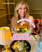 A bright, well-composed influencer-style shot featuring a creator presenting a plate of vegan fish and chips at a hotel restaurant. The image effectively uses depth of field to highlight the main dish while maintaining a clean, inviting atmosphere.