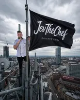 A promotional image featuring a private chef standing on a high-altitude structure overlooking the Manchester skyline, holding a branded flag. The image aims to convey high standards and an elevated experience, positioning the chef as a premium service provider for home dining.