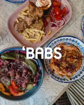 A high-quality, overhead flat-lay shot of three plates of Mediterranean-style food, including kebabs, fries, and hummus, featuring the restaurant name 'BABS' overlaid in the center.